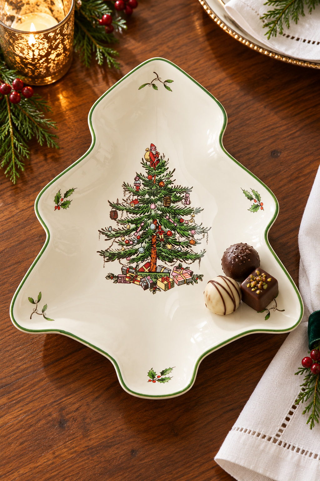 A tree-shaped cream serving dish with a green rim and printed decorated Christmas tree, holding a few chocolates on a wooden table.