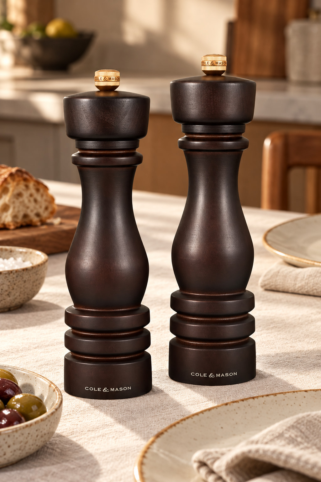 Two dark chocolate-coloured wooden salt and pepper mills with brass-coloured knobs, on a pale linen dining table.