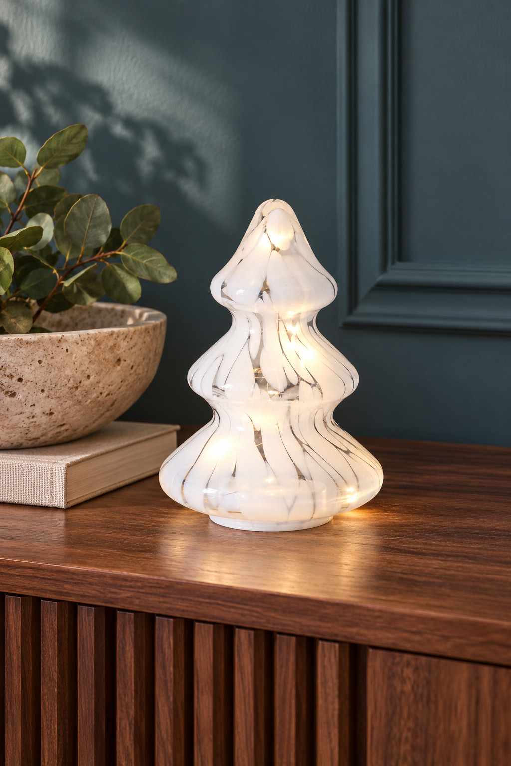 A small white marbled-glass Christmas tree lit from within, on a walnut cabinet against a deep teal wall.