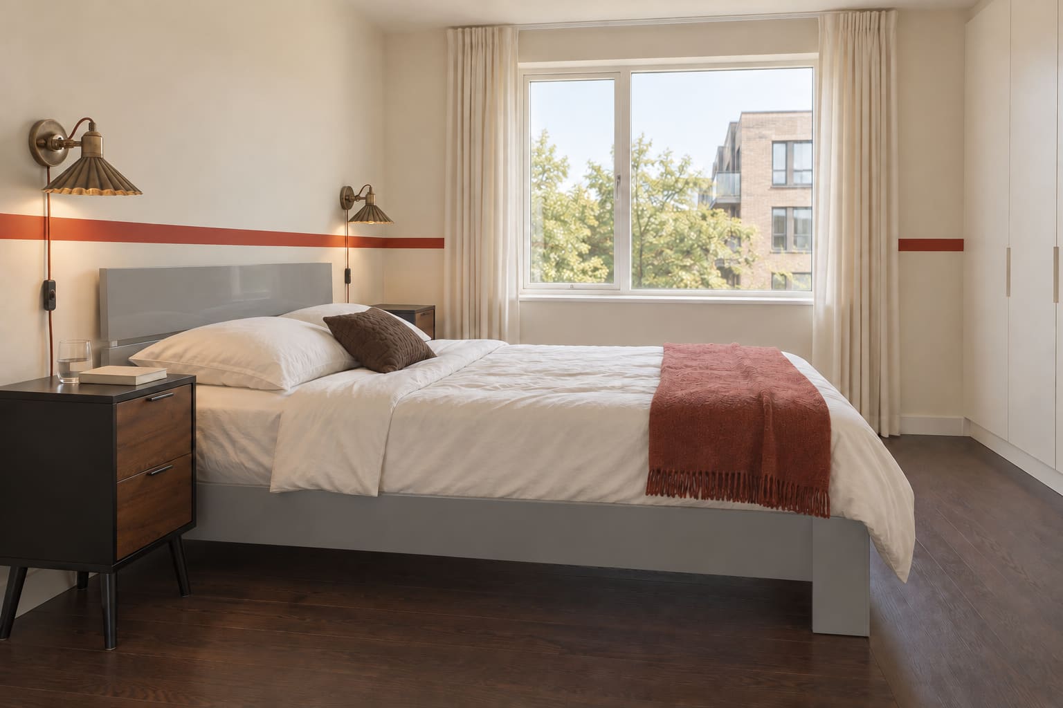 Side view of a cream apartment bedroom with one paprika-red wall stripe, a low grey lacquer bed, walnut-fronted black bedside cabinets, brass wall lamps and dark timber flooring.