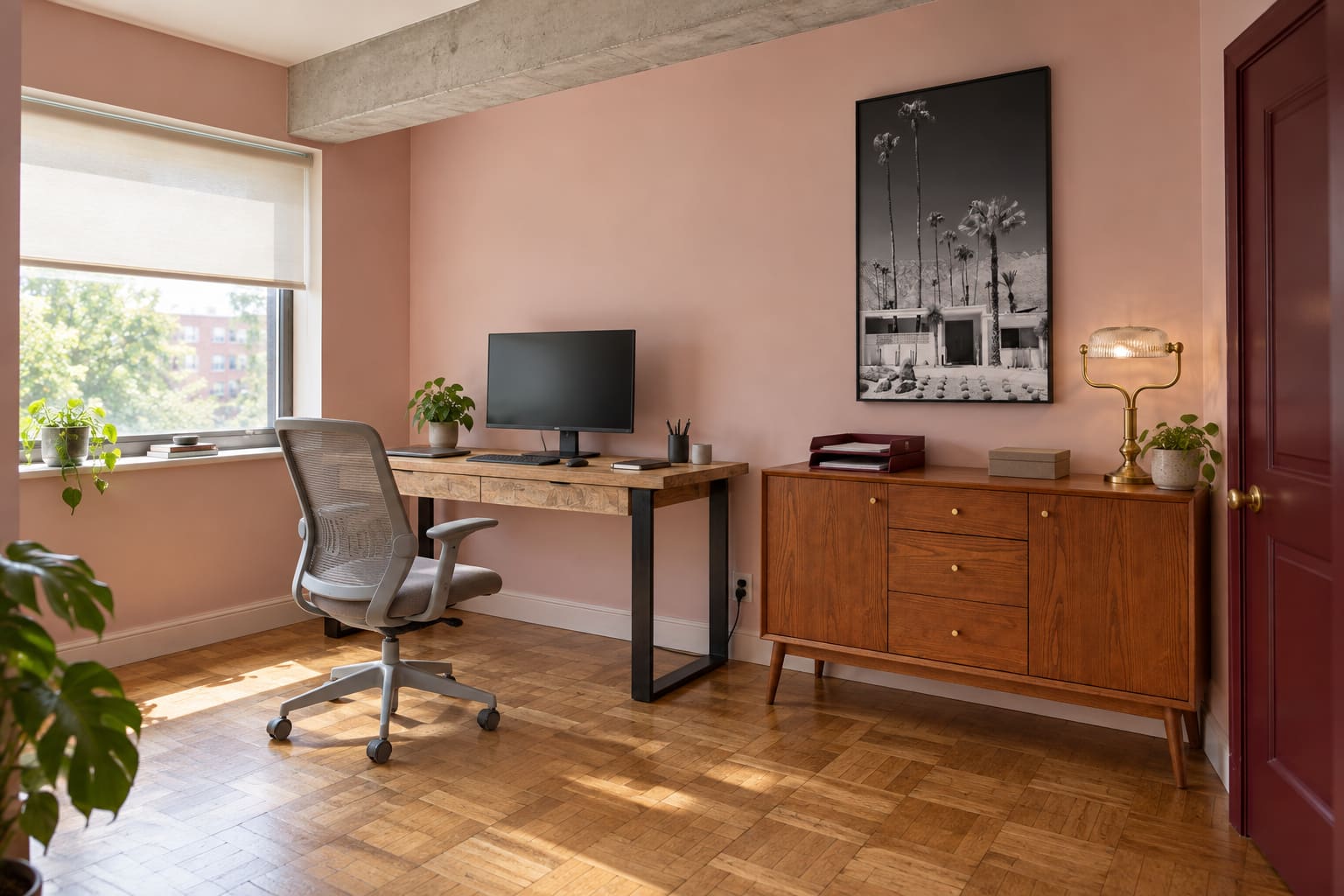 A blush-pink apartment office with an oxblood door, a mango-wood desk beside the window, a grey mesh office chair, cherry-ash storage and a black-and-white Palm Springs photograph.