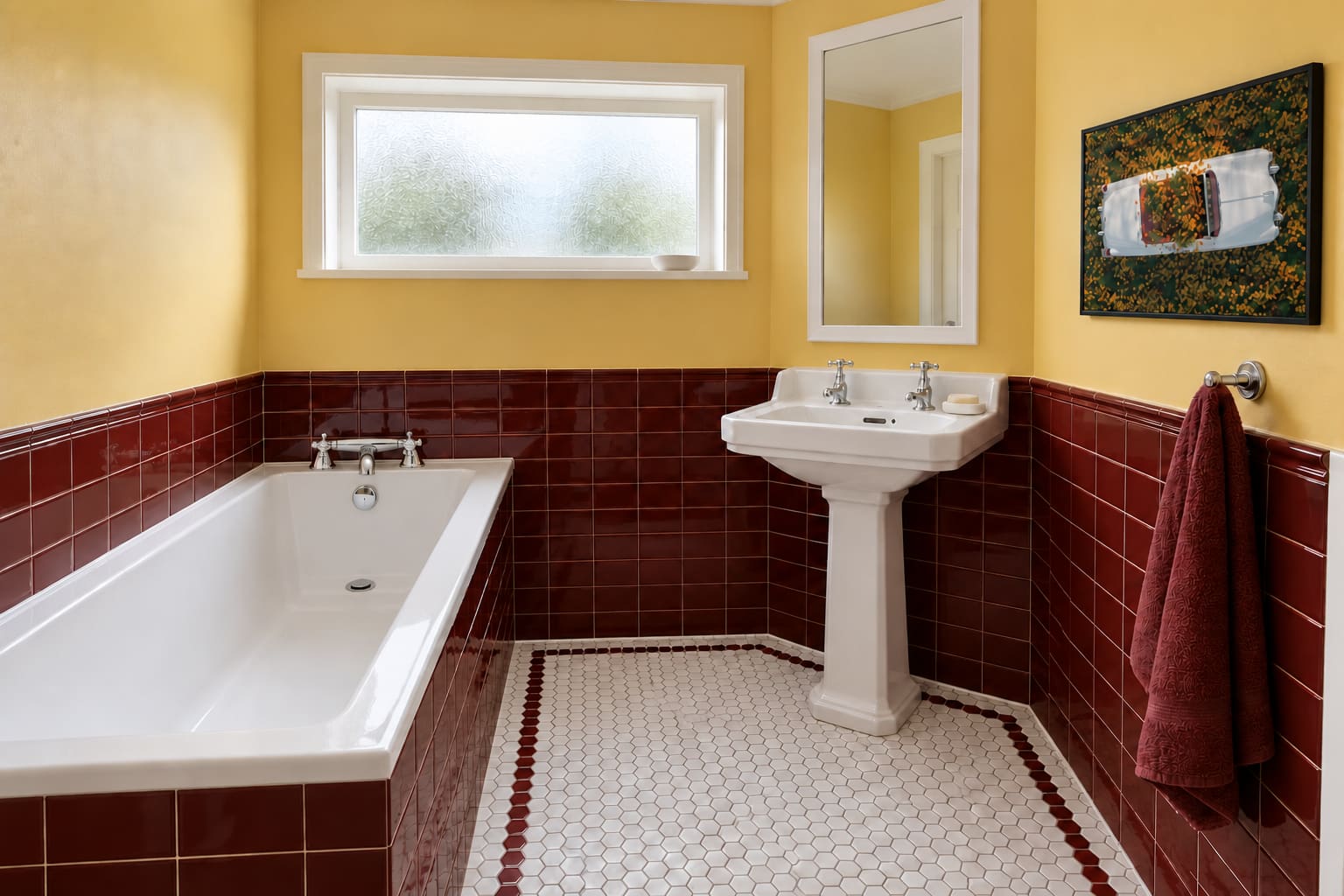 A butter-yellow bathroom with oxblood tiles, a white fitted bath, a traditional pedestal basin, a tall white-framed mirror and a photograph of a flower-filled convertible.