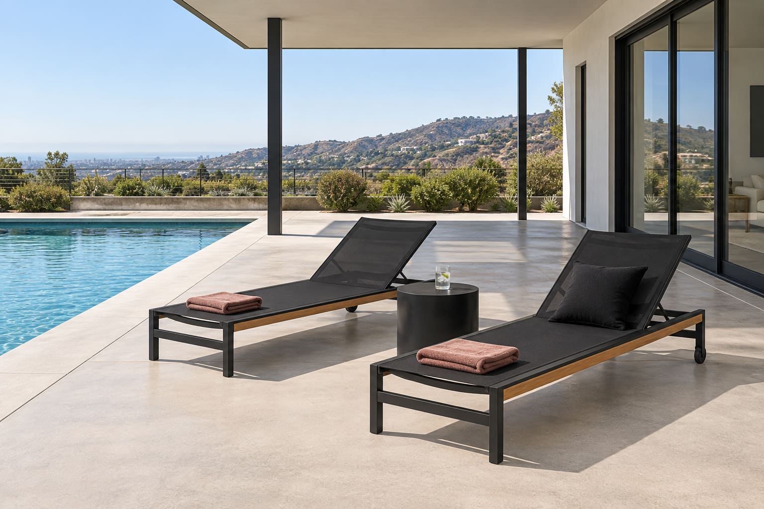 Level shaded pool terrace with two black mesh loungers, a black cylindrical table and rose towels overlooking dry hills.