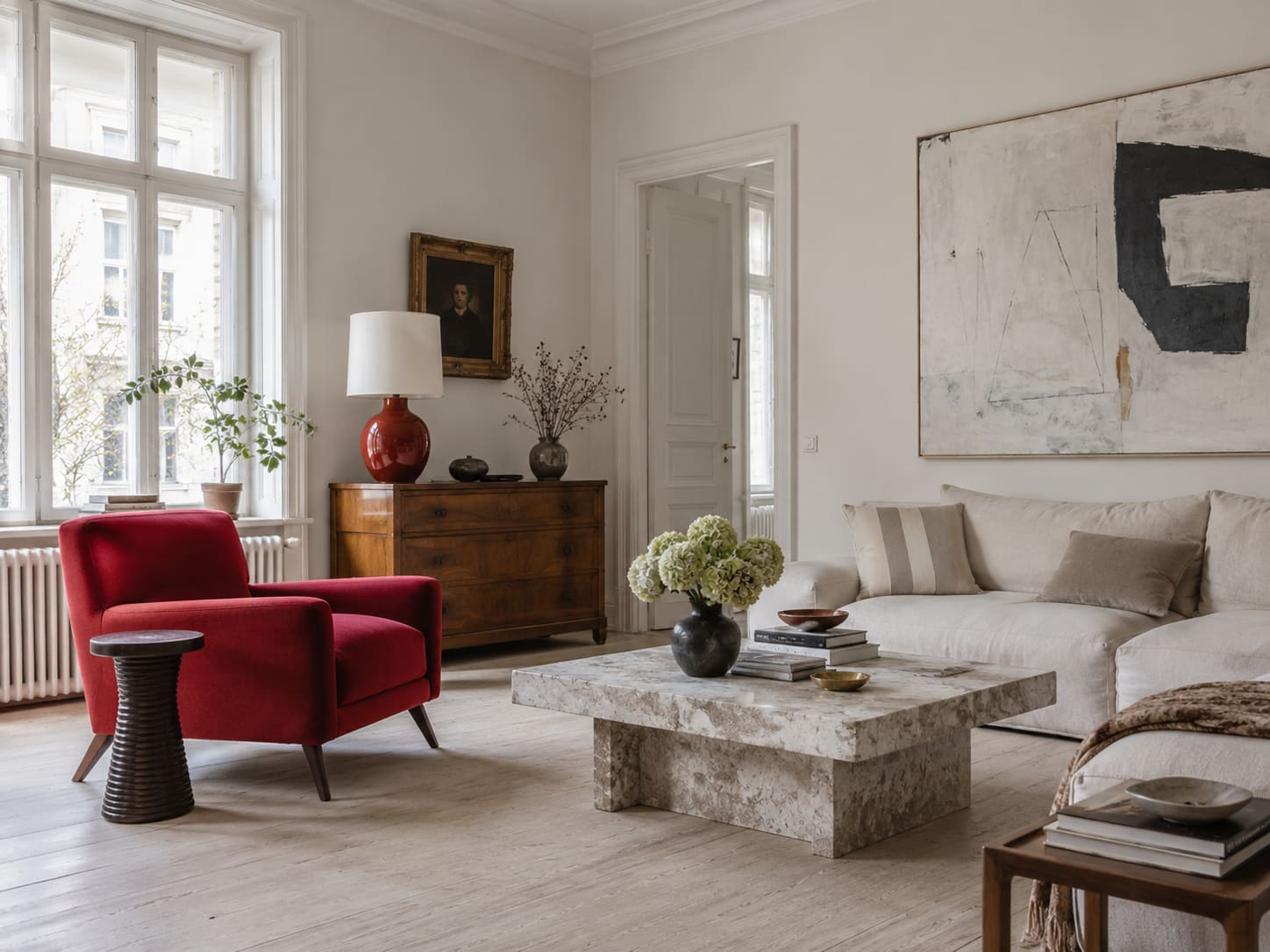 Red armchair and red ceramic lamp punctuating a pale living room with a timber chest and stone coffee table.