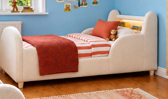 Pale upholstered single bed with orange striped bedding, a rust knitted throw and small personal pictures on a sky-blue wall.
