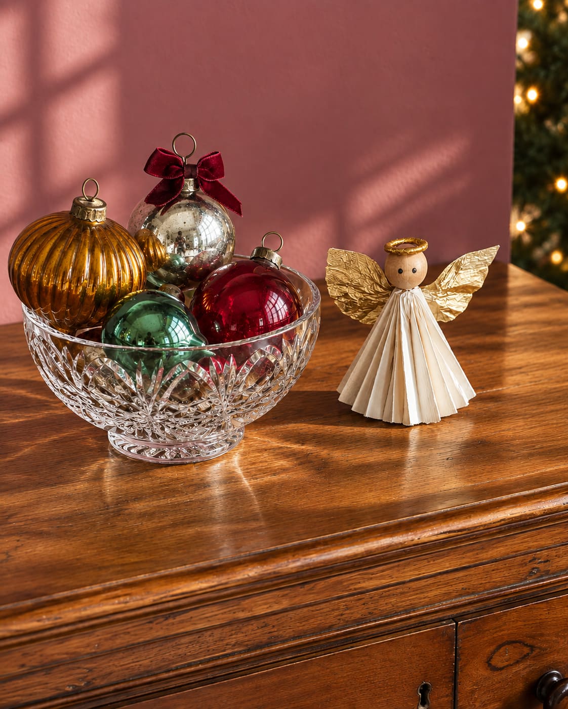 Red, green, amber and silver glass baubles in a cut-glass bowl beside a folded paper angel with gold wings, photographed in sunlight on a timber cabinet.